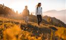 Two hikers enjoy nature at sunset. In the background, mountains and forests can be seen.