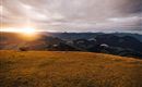 A beautiful mountain landscape at sunset, with several paragliders flying over the hills. The clouds are soft, and the colors of the sky are warm and inviting.