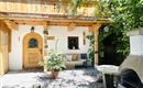 A cozy outdoor area with a wooden porch and plants. The entrance has a rustic door and there is a stone grill in the corner.