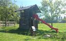 Een houten speelhuis met een rode glijbaan staat op een grasveld. Op de achtergrond zijn bomen en een hek te zien.