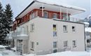 A modern building in the snow with white walls and red accents. The balcony is decorated with a snow overhang and there are some trees nearby.