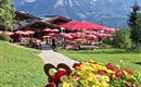 Een pittoresk restaurant met rode parasols en bloeiende bloemen op de voorgrond. Op de achtergrond verrijzen majestueuze bergen onder een heldere blauwe lucht.