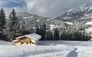 A snow-covered landscape with a cozy wooden house. In the background, snow-capped mountains and tall fir trees can be seen.