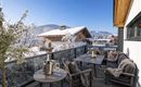 A cozy terrace with a table and chairs, surrounded by snow-capped mountains. In the background, traditional wooden houses and blue sky can be seen.