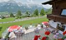A beautiful terrace with seating and red sun umbrellas. In the background, impressive mountains and a green landscape can be seen.