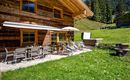 Eine gemütliche Terrasse mit einem Tisch und Liegestühlen, umgeben von grünem Gras und Bäumen. Im Hintergrund ist ein Holzhaus in einer malerischen Berglandschaft zu sehen. 
Chalet Heualm - Kelchsau Kitzbüheler Alpen