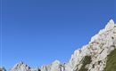An impressive mountain landscape with steep rocks and green terrain. The sky is clear and blue, while clouds rest in the valley.