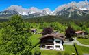 A picturesque landscape with a charming alpine chalet and green grass. In the background, majestic mountains rise under a clear blue sky.