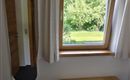 Een lichte kamer met een raam dat uitkijkt op een groene weide. Naast het raam staat een eenvoudige houten bank en een rechthoekige spiegel.