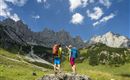 Two hikers are standing on a rock, gazing at majestic mountains. The sky is clear, and the landscape is green and vast.