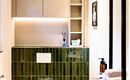 A modern bathroom with green tiles, an elegant toilet, and a sink. The walls are light, and there are practical shelves for storage.