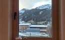 Ein Fenster mit Blick auf schneebedeckte Berge und ein typisches alpenländisches Dorf. Der Himmel ist leicht bewölkt und die Umgebung wirkt ruhig.