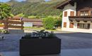 Eine gemütliche Terrasse mit einem Pflanzkübel und Blick auf grüne Hügel. Im Hintergrund sind traditionelle Häuser und ein klarer blauer Himmel zu sehen.