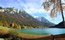 A tranquil landscape with a clear blue lake and majestic mountains in the background. The shore vegetation shines in vivid autumn colors.