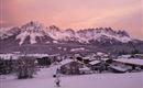 A picturesque winter landscape with snow-covered mountains and a pink sky. In the foreground, cozy houses can be seen in a snowy environment.