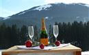 A bottle of sparkling wine is on a wooden table in the snow, surrounded by two glasses and red Christmas ornaments. In the background, snow-covered mountains and forests can be seen.