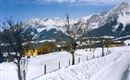 Eine winterliche Landschaft mit schneebedeckten Wiesen und Bergen im Hintergrund. Ein gelbes Haus steht am Rand der gefrorenen Straße.