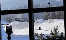 A view from a window of snow-covered roofs and snowy mountains in the background. The sky is clear and blue.
