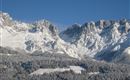 Snow-covered mountains under a clear blue sky. Below, a forest with freshly fallen snow.