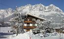 A charming wooden house in the snow, surrounded by snow-covered trees. In the background, majestic mountains rise under a clear sky.