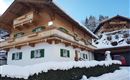 Ein traditionelles chaletartiges Haus im Schnee mit grünen Fensterläden. Die umliegende Landschaft ist winterlich und malerisch.