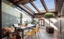 A bright, modern conservatory with a large dining table and comfortable chairs. Green plants and plenty of daylight create an inviting atmosphere.