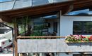 Ein schönes Holzhaus mit einer Terrasse, Wintergarten die mit Blumen dekoriert ist. Im Hintergrund sind Berge und ein blauer Himmel zu sehen.
