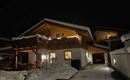 Een gezellig huis in de sneeuw, verlicht door warme lichten. De omgeving is kalm en winters.