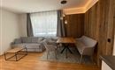 Ein modernes Wohnzimmer mit einem grauen Sofa, einem Holztisch und hellen Wänden. Die große Fensterfront lässt viel Licht herein und sorgt für eine freundliche Atmosphäre.