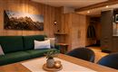 Ein modernes Wohnzimmer mit einer grünen Couch und einem Holztisch. Im Hintergrund sind Küchenschränke und ein schönes Bergbild an der Wand zu sehen.