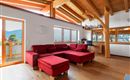 Ein modernes Wohnzimmer mit rotem Sofa und Holzbalkendecke. Große Fenster bieten einen Blick auf die umliegende Landschaft.<br/>Mit einer gemütlichen Bar integriert.