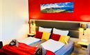Een moderne slaapkamer met een groot dubbelbed en kleurige kussens. Aan de muur hangt een landschapsschilderij, en de ruimte heeft een gezellige sfeer.