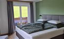 A modern bedroom with a large bed and green walls. Large windows let in plenty of daylight and offer a view of the outdoors.