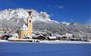 A picturesque winter landscape with snow-covered hills and charming cottages. In the foreground is a yellow church against a clear blue sky.