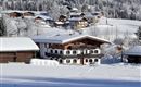 A snowy landscape with cozy wooden houses and snow-covered fields. In the background, more mountains and trees can be seen.