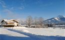 A winter landscape with snow-covered fields and mountains in the background. A cozy house stands in the foreground under a clear blue sky.
