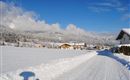 A snowy landscape with a clear blue sky. In the background, houses and mountains can be seen.