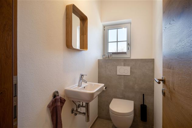 Ein modernes Badezimmer mit einem Waschbecken, einer Toilette und einem kleinen Fenster. Die Wände sind hell gestrichen und es gibt einen Holzrahmen für den Spiegel.