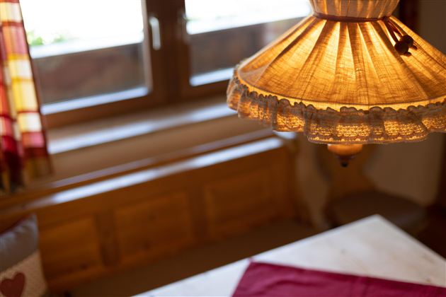 Eine gemütliche Stube mit einer warmen Lampe und einem Fenster im Hintergrund. Auf dem Tisch liegt ein rotes Tuch.