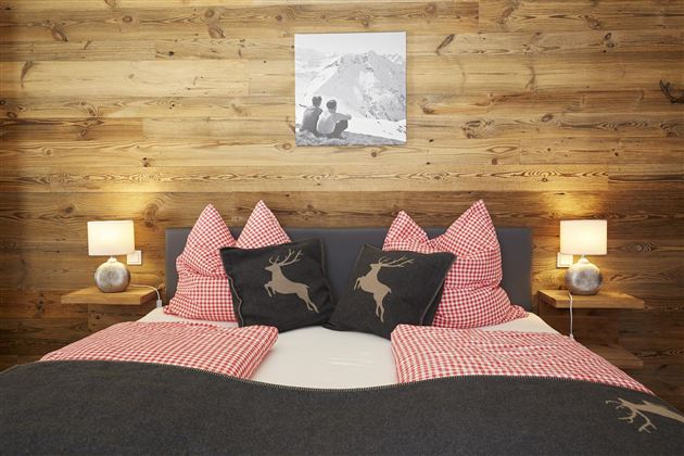 Ein gemütliches Schlafzimmer mit einem Holzwand und einem großen Bett. Die Kissen sind rot-weiß kariert und zeigen Hirschmotive, dazu gibt es warme Lampen.