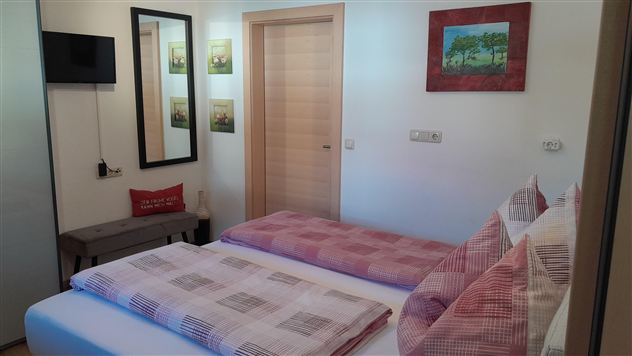 A cozy bedroom with two beds and red pillows. On the wall hang pictures and a mirror, there is a small television and a wooden door.
