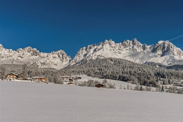 A picturesque winter landscape with snow-covered mountains and clear blue sky. In the foreground, there are traditional cottages in a tranquil setting.