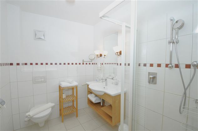 A modern bathroom with white tiles and a clear shower area. There is a wooden shelf with towels and a stylish sink setup.