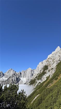 An impressive mountain landscape with steep rocks and green terrain. The sky is clear and blue, while clouds rest in the valley.
