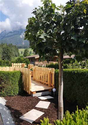 Eine stilvolle Holzbrücke führt durch eine grüne Heckenlandschaft. Im Hintergrund sind Berge und ein blauer Himmel zu sehen.