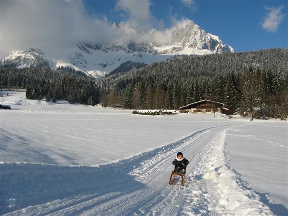 A boy is riding a sled on a snowy field. In the background, there are mountains and a wooden house.