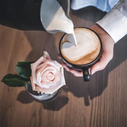 Een hand giet melk in een kopje koffie. Naast de kop staat een mooie roze roos.