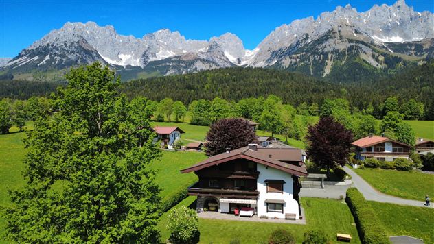 A picturesque landscape with a charming alpine chalet and green grass. In the background, majestic mountains rise under a clear blue sky.