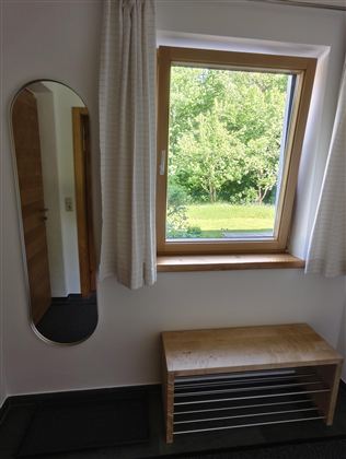 Een lichte kamer met een raam dat uitkijkt op een groene weide. Naast het raam staat een eenvoudige houten bank en een rechthoekige spiegel.
