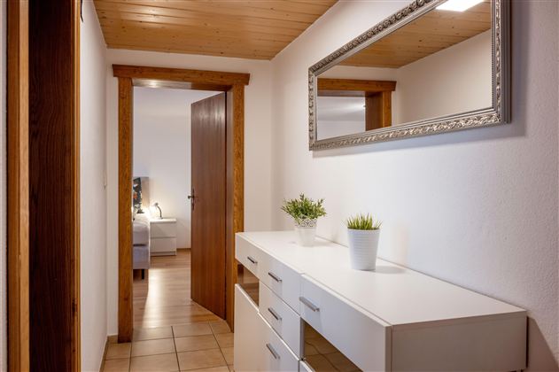 Ein heller Flur mit Holzdecke und einer weißen Kommode. An der Wand hängt ein Spiegel, und eine Pflanze steht auf der Kommode.